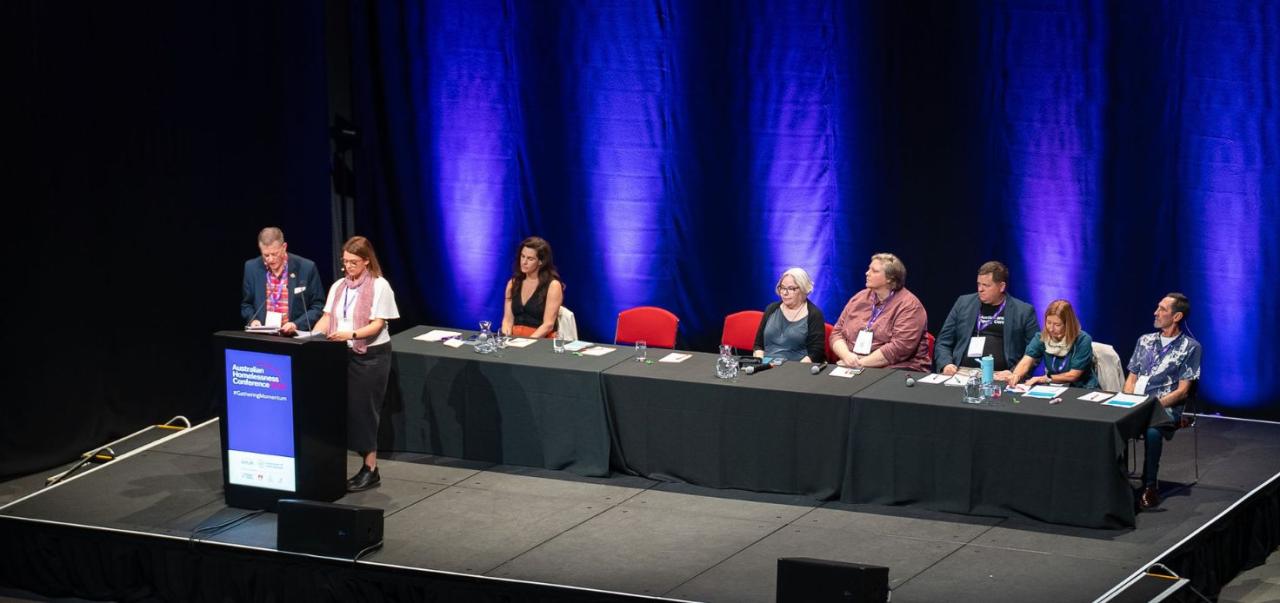 Australian Homelessness Conference 2024 major concurrent session. Two speakers are standing at the lectern and six are seated behind a long table on the stage.