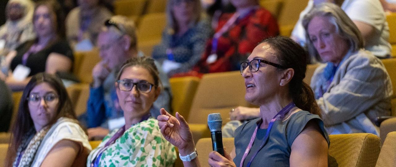 Close up of 3 women seated in an auditorium during conference Q&A. One woman is holding a microphone and speaking.