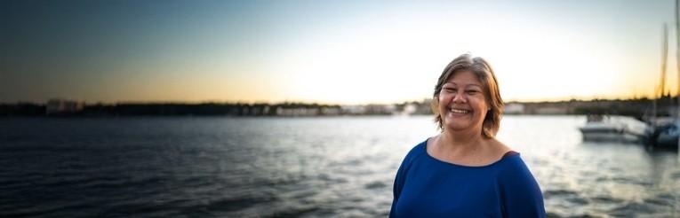 Middle aged woman standing with her back to a lake at sunset, smiling toward the camera
